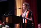 Woman in formal graduation regalia giving a speech at a lectern.